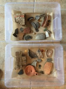 A plastic box containing layers of broken pieces of pottery, jewellery, mosaics on a layer of sand.
