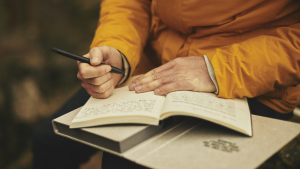 Hand holding a pen ready to write in a notebook. The book rests on a larger book, possibly on the writer's knee.