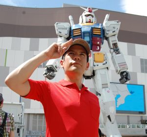 Daniel Fandino, a PhD Candidate in History at MSU, salutes the camera while standing in front of a towering Gundam, a giant robotic mech.