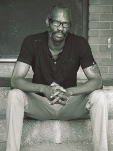 Cartoonist Ho Che Anderson sits on the cement steps of a building in a black short and khaki pants.
