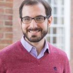 A man stands for a portrait-style photo; the brick building behind him is blurred. He smiles at the camera with brown eyes behind black glasses. He has white seeming skin, short brown hair and brown facial hair. He wears a red sweater over a purple button-up shirt.