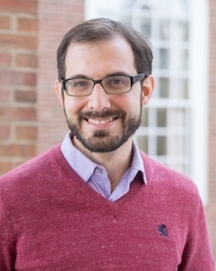 A man stands for a portrait-style photo; the brick building behind him is blurred. He smiles at the camera with brown eyes behind black glasses. He has white seeming skin, short brown hair and brown facial hair. He wears a red sweater over a purple button-up shirt.