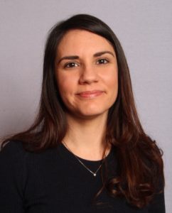 A woman stands in front of a white background, smiling at the camera with brown eyes. She has tan seeming skin, brown hair down past the shoulder, and is wearing a black top.