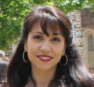 A woman stands in front of a building, smiling at the camera with brown eyes. She has long brown hair and light brown seeming skin. She is wearing a brown top, gold hoop earrings, and red lipstick.