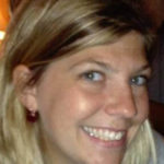 A woman smiles at the camera with blue eyes. She has blonde hair with brown highlights and white seeming skin.She is wearing a red and white striped top and red earrings.