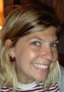 A woman smiles at the camera with blue eyes. She has blonde hair with brown highlights and white seeming skin.She is wearing a red and white striped top and red earrings.