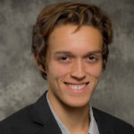 A white man with brown hair is seated in front of a grey background. The man wears a black suit jacket and a grey, buttoned shirt.