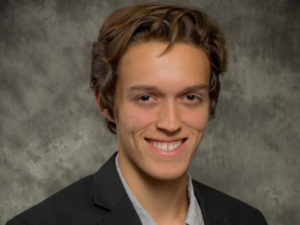 A white man with brown hair is seated in front of a grey background. The man wears a black suit jacket and a grey, buttoned shirt.