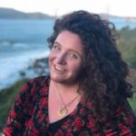 A white woman sits on a shoreside hill with the Golden Gate Bridge blurry in the background. The woman wears a soft black and red velvet dress and a gold sunburst necklace. She has a purple wheelchair and a lot of red hair.