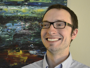 A smiling man stands in front of an abstract painting that has streaks of red, black, green, and blue. He has short brown hair and is wearing dark glasses and a blue button-up shirt.