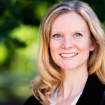 A woman stands for a portrait-style photo with blurred greenery in the background. She smiles at the camera with blue eyes. She has shoulder-length blonde hair and white seeming skin. She is wearing a black blazer over a white top and silver stud earrings.
