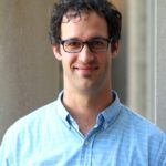 A man stands for a portrait-style photograph with blurred columns behind him. He has curly black hair and smiles at the camera with brown eyes behind blue glasses. He is wearing a blue and white striped button up shirt.