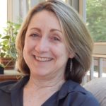 A woman sits on a couch in front of a window. She smiles at the camera with green eyes. She has shoulder length blonde hair and white seeming skin. She is wearing a blue blouse and silver wire earrings.
