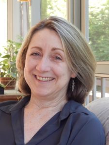 A woman sits on a couch in front of a window. She smiles at the camera with green eyes. She has shoulder length blonde hair and white seeming skin. She is wearing a blue blouse and silver wire earrings.