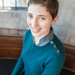 A woman sits for a portrait-style photograph. She has white seeming skin and looks into the camera with blue eyes, smiling. She is wearing a blue sweater over a blue shirt and silver stud earrings.