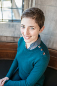 A woman sits for a portrait-style photograph. She has white seeming skin and looks into the camera with blue eyes, smiling. She is wearing a blue sweater over a blue shirt and silver stud earrings.