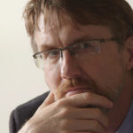 A man looks closely at the camera with brown eyes behind glasses and a hand on his chin. He has short dark brown hair, light brown facial hair, and white seeming skin. He is wearing a charcoal suit over a blue shirt.