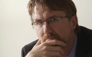 A man looks closely at the camera with brown eyes behind glasses and a hand on his chin. He has short dark brown hair, light brown facial hair, and white seeming skin. He is wearing a charcoal suit over a blue shirt.