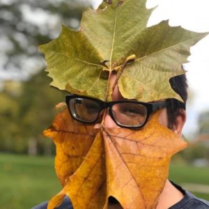 A man stands in front of an autumnal landscape. His face is obscured by two very large leaves: an orange leaf covers his mouth and chin and a green leaf covers his forehead. In between the leaves can be seen a white nose and brown eyes behind thick black glasses.