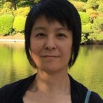 A woman stands in front of a lake and trees, smiling at the camera. She has straight black hair, light brown seeming skin, and brown eyes. She wears a black top with a black cardigan and a silver necklace