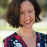 A woman looks into the camera with a blurred, natural background. She has white skin, brown hair, and is wearing a top that has splashes of white, blue, black, and red.