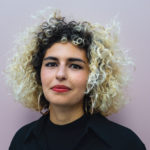 A woman with brown eyes and honey-colored skin looks into the camera intensely. She has curly blonde hair with brown roots. She wears gold hoop earrings and a black top.