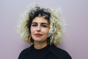 A woman with brown eyes and honey-colored skin looks into the camera intensely. She has curly blonde hair with brown roots. She wears gold hoop earrings and a black top.