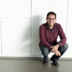 Ronen Steinberg sitting against a wall in the Broad Museum at Michigan State University, as photographed by Jackie Hawthorne. He wears a red shirt and glasses and has dark hair.
