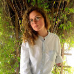 A woman stands in front of a plant leaning on a trellace. She looks at the camera with confidence, not smiling. She has green eyes behind tortiseshell glasses and shoulder length light brown hair and white seeming skin. She wears a blue button-up shirt with one sleeve rolled up to show an arm that ends around the elbow