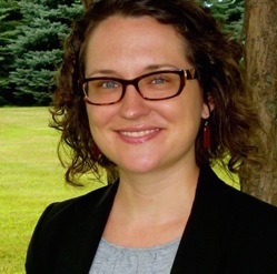 A woman stands in front of a field and pine trees. She smiles at the camera with blue eyes behind tortoiseshell glasses. She has shoulder-length curly brown hair and white seeming skin. She is wearing a black blazer over a grey top and dark red earrings.