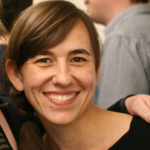 A woman with brown hair in a ponytail down to her shoulder and white seeming skin leans against the shoulder of another person outside of the frame. She has brown eyes and wears a black top.