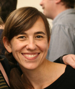 A woman with brown hair in a ponytail down to her shoulder and white seeming skin leans against the shoulder of another person outside of the frame. She has brown eyes and wears a black top.