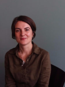 A woman sits for a professional headshot smiling at the camera. She has white seeming skin, brown hair, brown eyes, and is wearing a brown button-up shirt and a silver necklace.