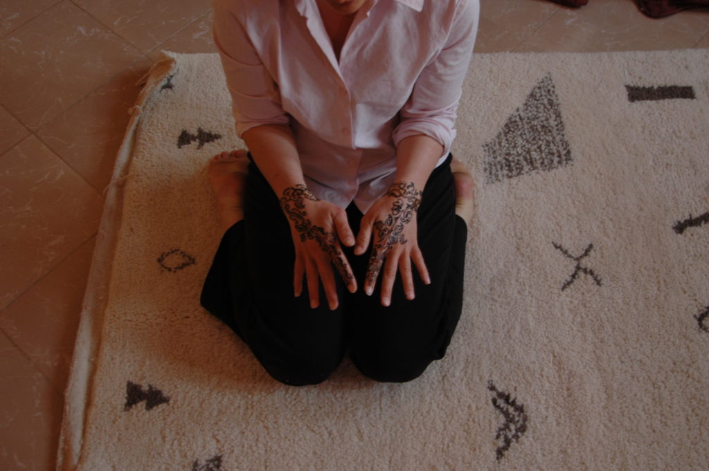 Image shows a woman on a rug with patterns on it and just shows her hands which are decorated with henna.