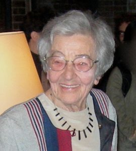 Cropped picture of Ursula Franklin, a white woman with grey hair and glasses, in front of a lamp wearing a grey shirt with red and blue stripes of varied sizes and a necklace with multiple rectangles.