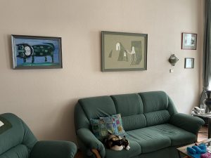 Buttercup, a calico cat is curled up on a green sofa. On the beige wall above her are two large paintings, one of them depicting a silly cat, an artful wall sconce, and two small paintings.