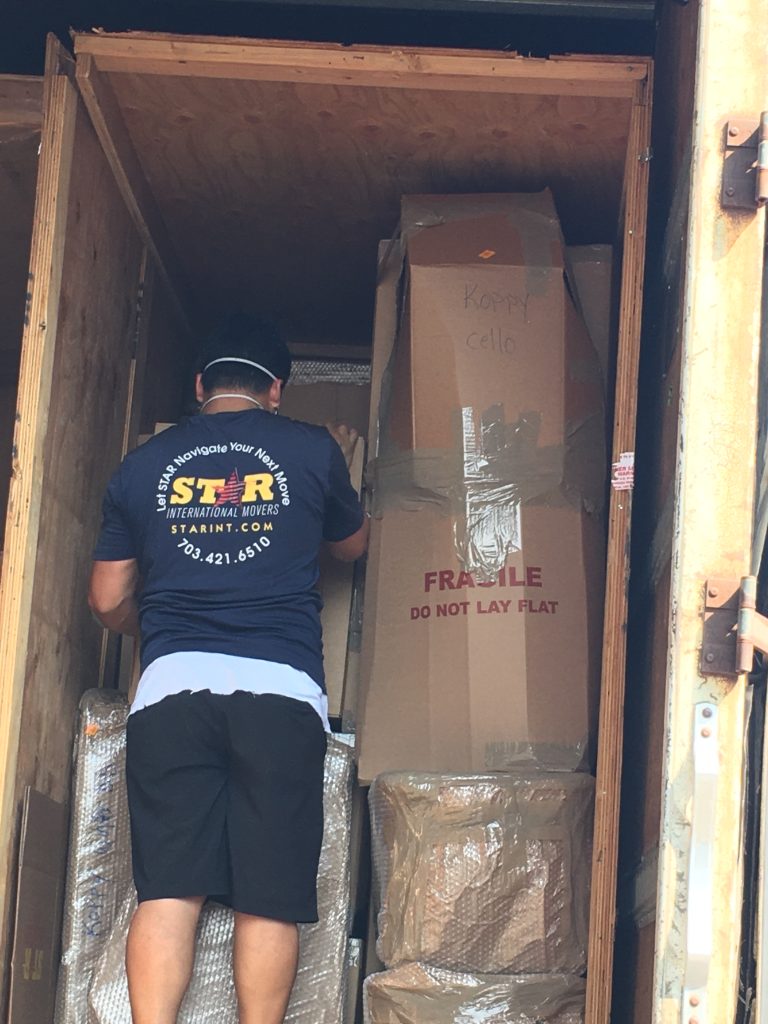 A shipping crate half full of cardboard boxes. The crate is twice as wide as the mover standing in front of it and a third again taller.