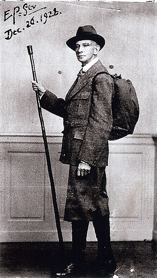 Photograph of Edward Prime-Stevenson, a white man standing and wearing a hat, long dark-colored coat, boots, and carrying a backpack and long walking stick. The upper left-hand corner is signed "E.P. St., Dec. 26, 1928."